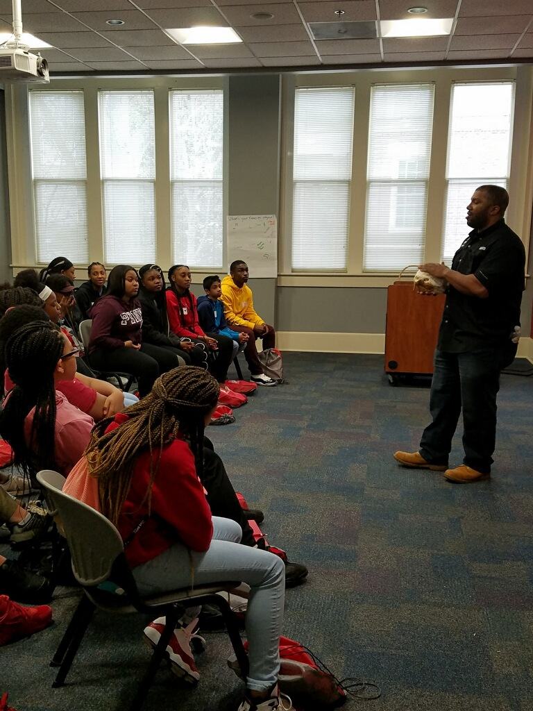 Dr. Newberry III stands at the front of a classroom addressing a group of young students