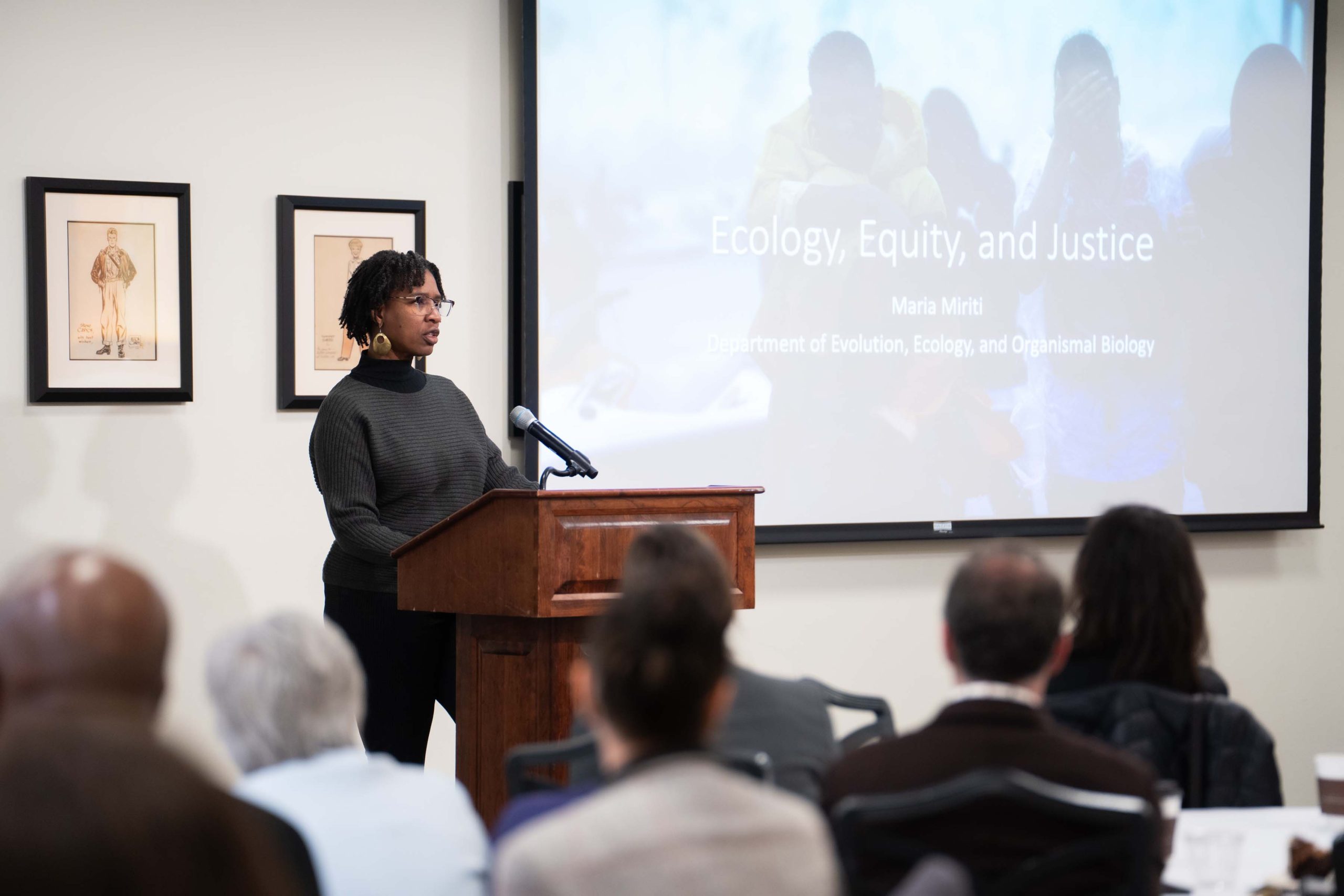 Dr. Maria Miriti is pictured speaking behind a podium next to a projected title slide which reads: Ecology, Equity, and Justice