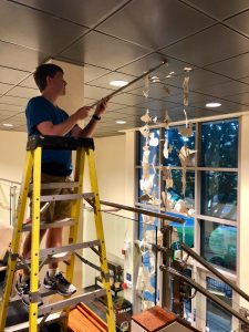 Man on a step ladder, hanging artwork from the ceiling. Artwork looks like a mobile.