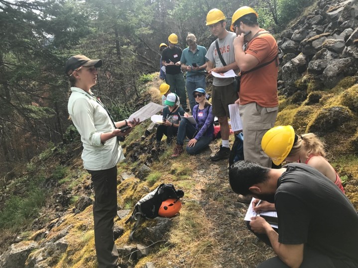 Woman (left) standing and explaining to group (right; on trail). Nearly all group members are wearing yellow hard hats. Group is in a forest, on a trail.