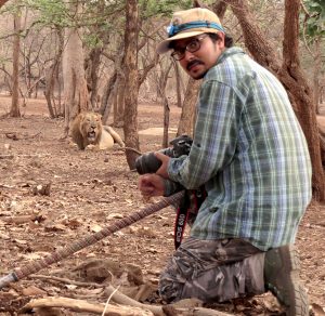 The author, wearing a baseball hat, glasses, a plaid shirt, and camouflage pants, holding a camera and looking back at the photographer. He is crouched, and in the background there are trees and a male lion.