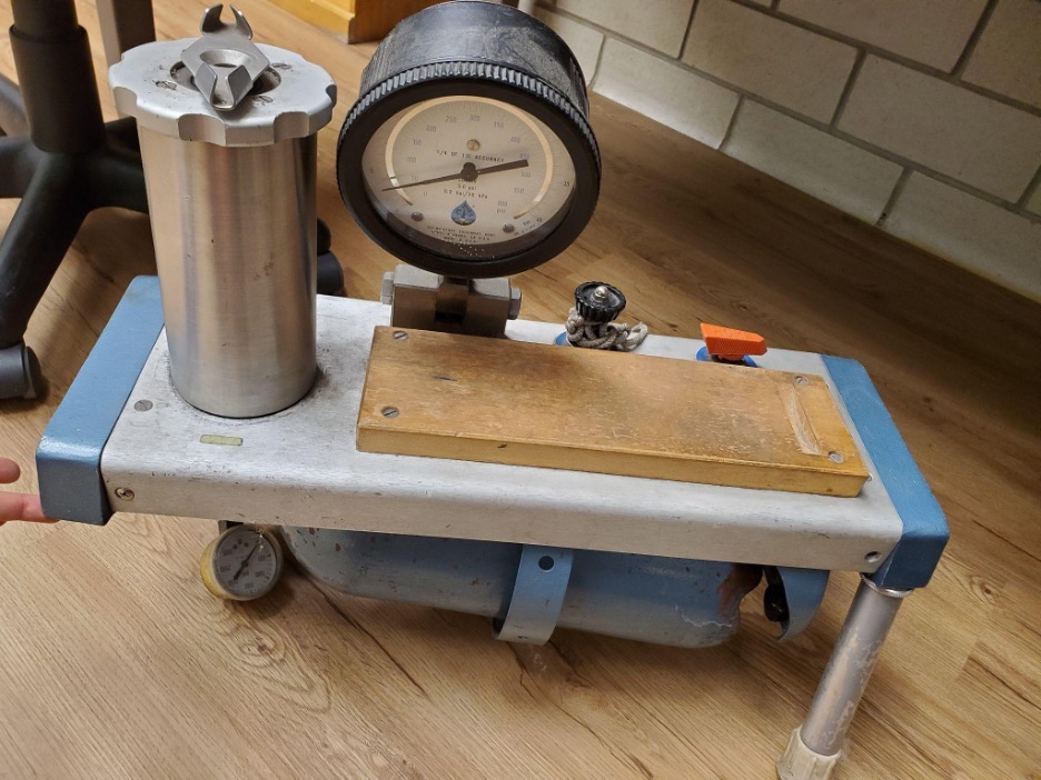 Title: The Multi-Decade-Old, Not-Quite-Functioning, 3-Legged Pressure Chamber The image shows a metal contraption called a Scholander Pressure Chamber. It sits on a wooden floor in front of a table and rolling chair. The Chamber has a nitrogen gas tank strapped to the bottom, a small pressure meter to the left of the gas tank, a larger pressure meter on the top, a metal cylinder to the upper left, and two valves to the right of the pressure meter. It is missing its front right leg, so it only stands upright with the support of my finger.