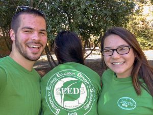 Three students in green shirts, one facing backwards to show their SEEDS shirt graphic