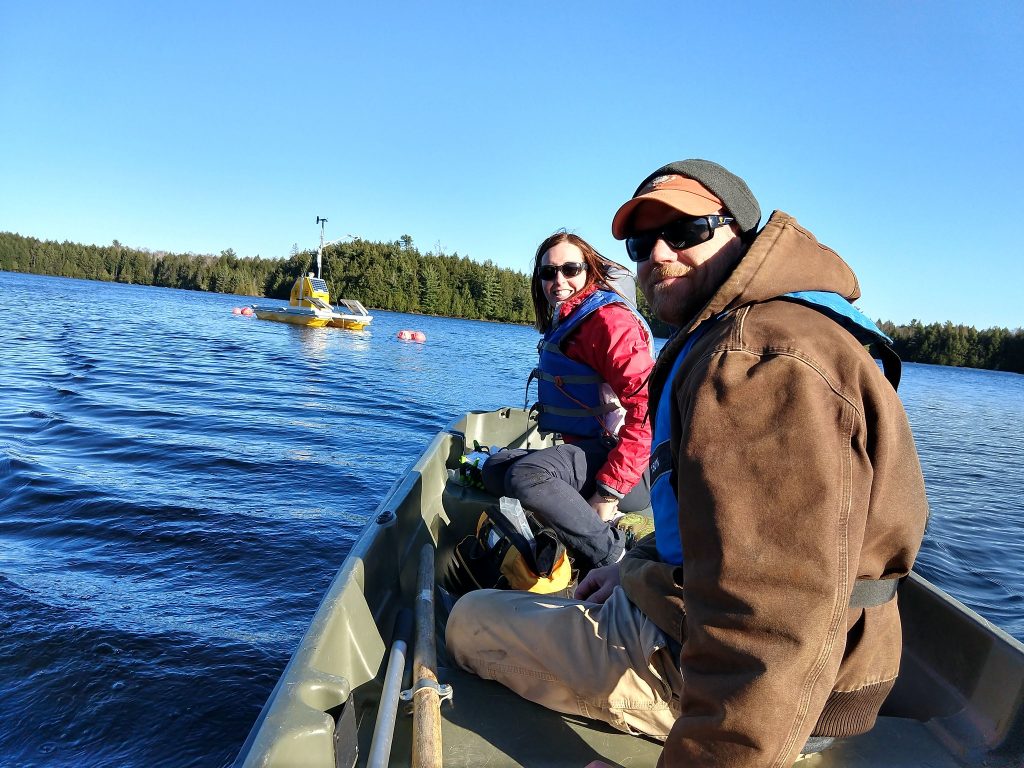  Two field technicians sit in a small raft or boat, looking back at the camera. Both are wearing coats and life preservers and have a bag full of gear between them. They are approaching a data collection station in the middle of a lake lined with evergreen trees. The data station is yellow and is fitted with several solar panels. 