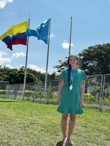 A person standing outdoors in a green dress, smiling in front of three flagpoles with colorful flags, at COP16 in Cali, Colombia, on a sunny day.