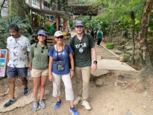 Three hikers pose together on a forest trail in front of a juice stand, surrounded by greenery.