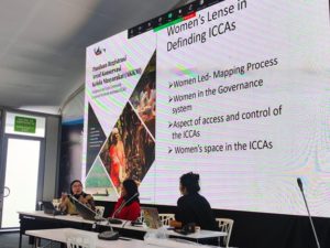 Cindy Julianty presenting during a side event at COP16, sitting alongside two panelists with a large screen displaying a presentation on "Women's Lens in Defining ICCAs".