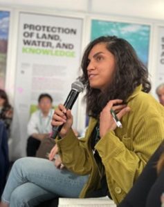 A woman speaking into a microphone during a conference session at COP16, with a focused expression.