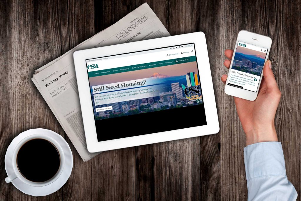 A newspaper sits under a tablet next to a cup of coffee. An arm is outstretched with a cell phone which shows the same ESA homepage as the tablet.