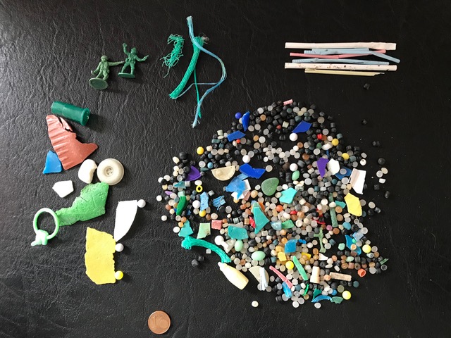 A cluster of small, colorful plastic pellets, broken shards, and small toys on a black leather surface. There is a 1-cent coin at the bottom of the photo for scale.