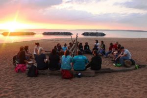 Image of a group surrounding a bonfire on a beach with the sun setting.