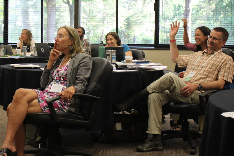 A group of participants in a lecture smile some with raised hands.