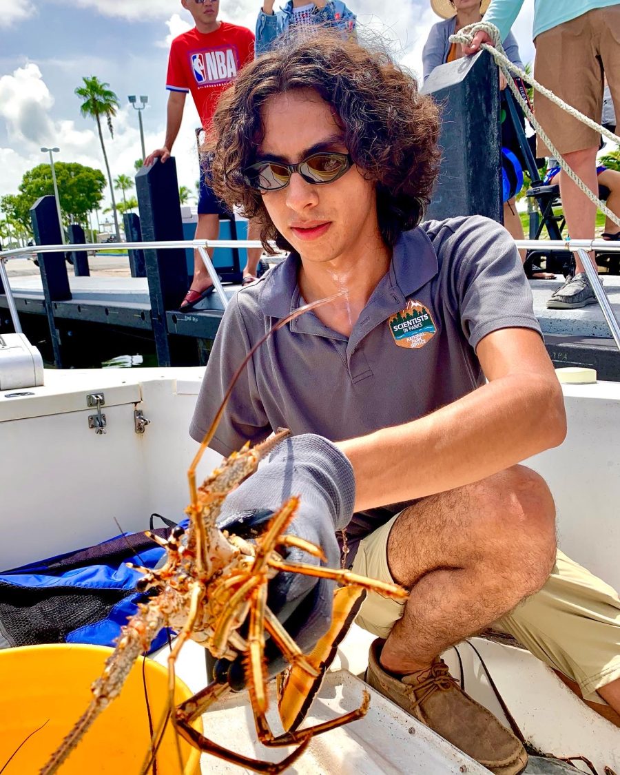 Student squats on a boat and holds a lobster in his hand as people watch behind him. 