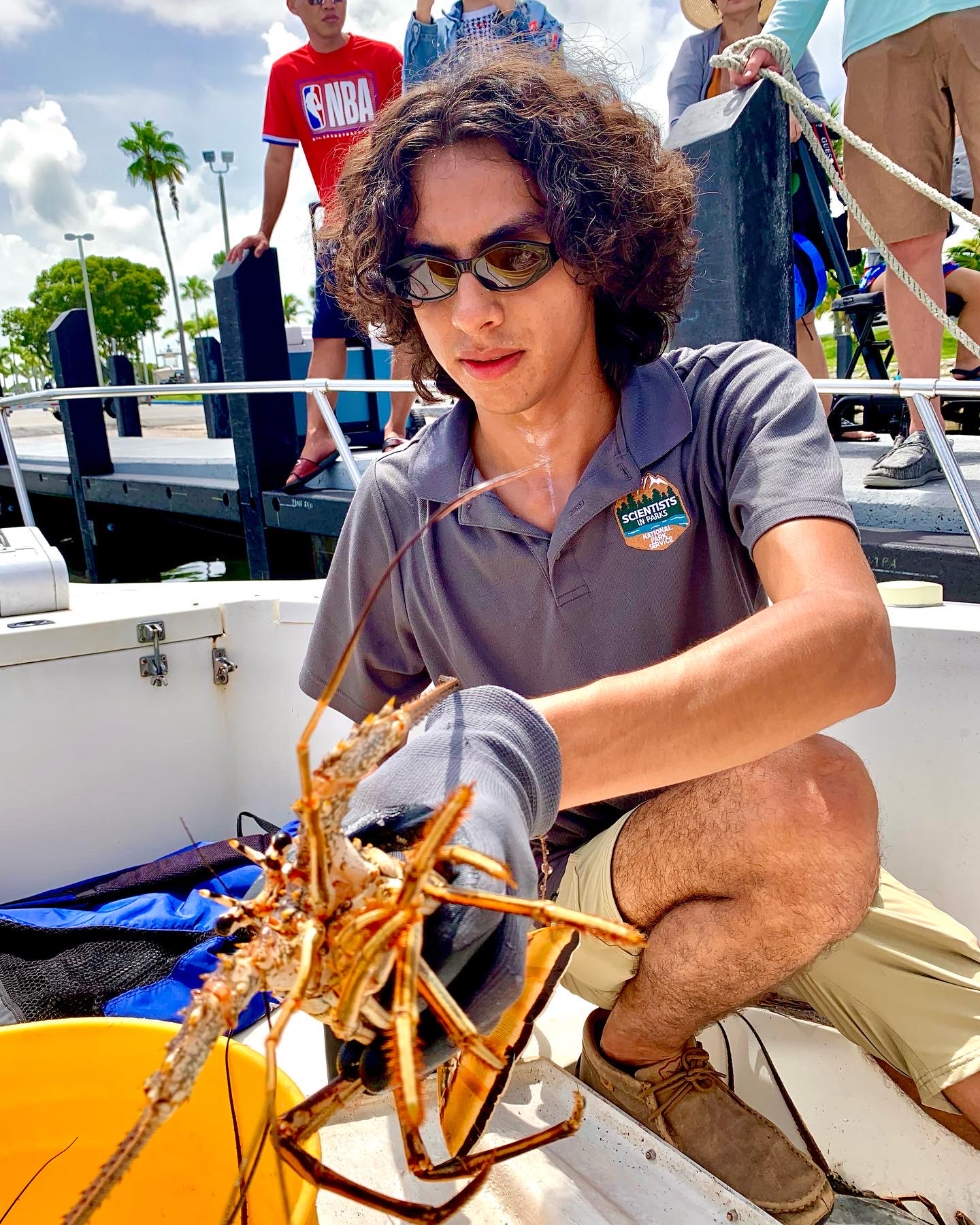 Student squats on a boat and holds a lobster in his hand as people watch behind him.